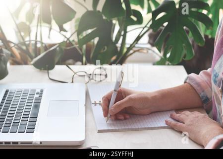 Mani di una donna seduta alla scrivania di fronte al computer portatile che scrive appunti Foto Stock