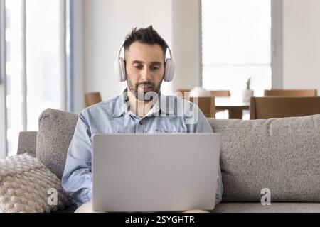 Uomo ispanico seduto sul divano e con le cuffie sul computer portatile Foto Stock