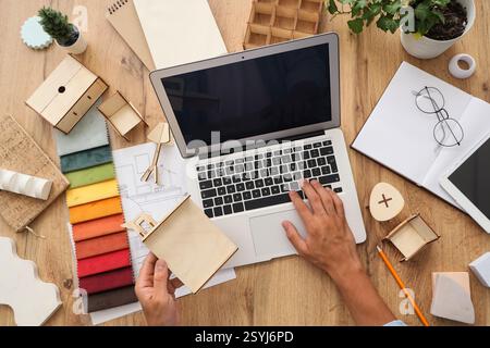 Progettista di mobili maschile che lavora con un computer portatile e un modello in miniatura su un tavolo in legno, vista dall'alto Foto Stock