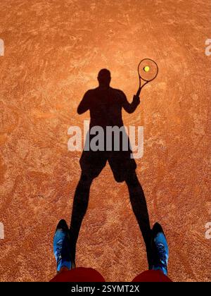 Uomo che gioca a tennis in una giornata di sole. Guardando in basso l'ombra del tennista su un campo di argilla sabbiosa. Foto Stock