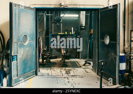 Grandi porte industriali blu si aprono per rivelare uno spazio interno con apparecchiature, cavi e strutture metalliche in fabbrica. Foto Stock