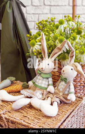 Due coniglietti di peluche fatti a mano, conigli con carote decorative sulla superficie in vimini sullo sfondo del bouquet di fiori. Concetto di vacanza di Pasqua Foto Stock