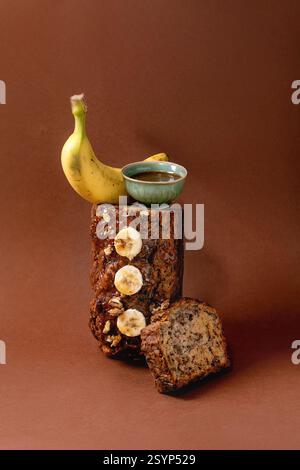 Natura morta moderna con pane alla banana fatto in casa. Pagnotto con noci, caramello gocciolato, banana fresca ricoperta su fondo marrone. Banana e salsa Foto Stock