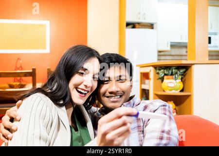 Una coppia gioiosa si abbraccia sul divano, sorridendo mentre guardano un test di gravidanza positivo. condividono un momento di cuore, celebrando l'anticipazione di Foto Stock