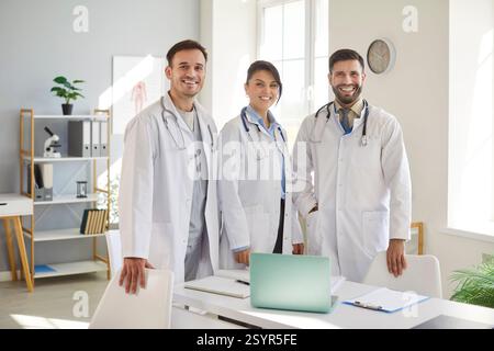 Felice team di medici professionisti in cappotti bianchi in piedi in ufficio medico e sorridendo alla macchina fotografica. Foto Stock