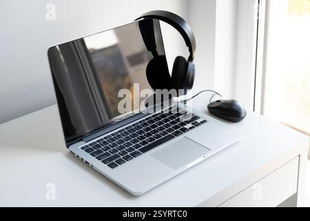 Laptop aperto color argento, cuffie wireless nere e mouse nero per computer su una scrivania bianca Foto Stock