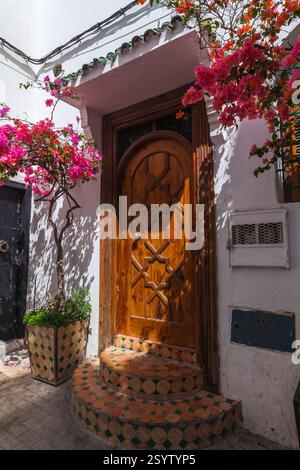 Ingresso ad un tradizionale edificio marocchino, caratterizzato da una porta di legno splendidamente intagliata fiancheggiata da vibranti bouganville, che creano un'affascinante miscela Foto Stock