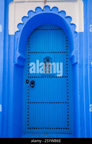 porta in legno ad arco blu con borchie decorative e una piccola finestra ornata, adagiata su una parete blu e bianca Foto Stock