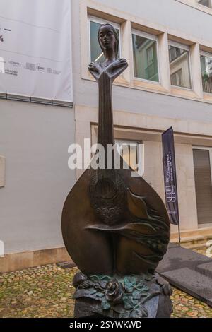 Una scultura in bronzo di una figura femminile che si fonde con un liuto sorge in un cortile lastricato contro un edificio moderno Foto Stock