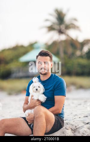 Giovane con il suo piccolo cane bianco maltese sulla spiaggia al tramonto Foto Stock