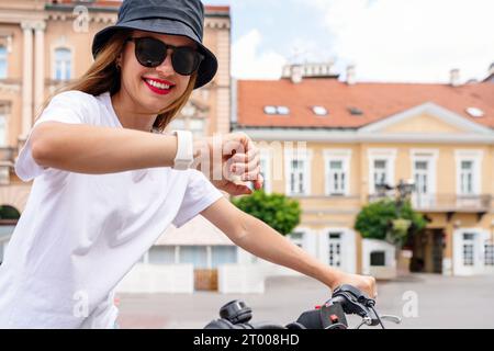 Giovane donna urbana che guarda l'orologio al polso mentre va in bicicletta in città. Foto Stock