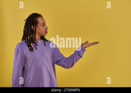 uomo afro-americano confuso in felpa viola con dreadlock che tiene la mano vuota con il palmo in alto Foto Stock