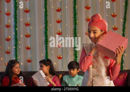 Ragazzo Sikh in piedi con un regalo in occasione di Diwali e dei suoi amici seduti dietro Foto Stock