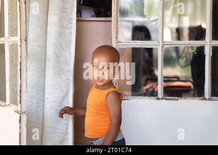 bambino africano di fronte alla casa nella township, che indossa un giubbotto arancione, in piedi davanti alla porta Foto Stock