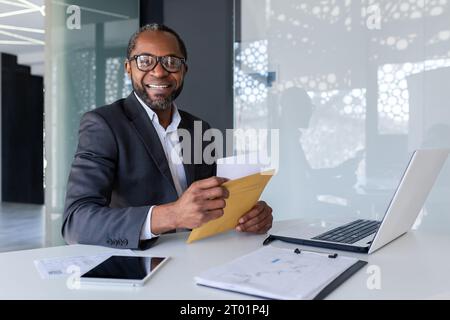 Ritratto di un giovane uomo d'affari afro-americano che lavora in ufficio a un portatile, tenendo in mano e aprendo una lettera ricevuta. Sorridere alla telecamera. Foto Stock