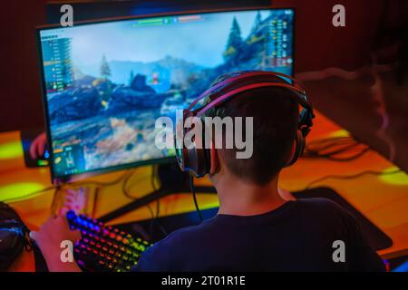 Vista posteriore di un uomo irriconoscibile in abiti casual indossando le cuffie mentre si gioca a un videogame per computer al tavolo Foto Stock