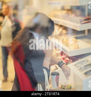 Donna irriconoscibile che chiama dal cellulare, che si prende una tazza di caffè da asporto e un'insalata. Pausa ufficio. Una vera e propria catena di alimentari. Vista sfocata in vetro Foto Stock