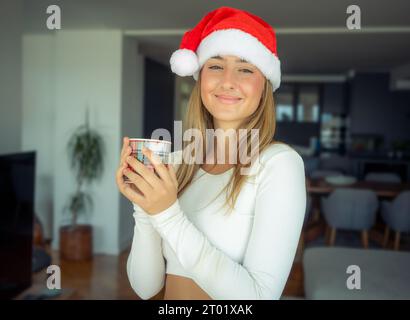 Giovane donna che indossa il cappello di babbo natale che beve caffè a casa Foto Stock