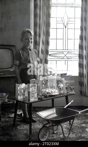 Anni '1960, storico, all'interno di una stanza, di fronte a una televisione dell'epoca, un bambino di 6 anni con orgoglio in piedi con la sua torta di compleanno e i biglietti di compleanno su un tavolo e uno dei suoi regali, una piccola carriola di metallo, Inghilterra, Regno Unito. Foto Stock