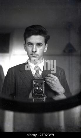 Anni '1950, storico, un giovane ma in una camicia e cravatta con una macchina fotografica pieghevole, forse una macchina fotografica pieghevole Ihagee del 1939, che porta il suo autoritratto in uno specchio. Foto Stock