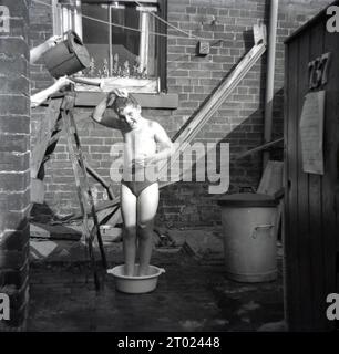 Anni '1960, storica, doccia fai da te..... In un cortile sul retro, un ragazzo con i suoi costumi da bagno in piedi in una ciotola di plastica che gli è stata versata dell'acqua da un annaffiatoio può essere tenuto da una persona su una scala, Oldham, Manchester, Inghilterra, Regno Unito. Foto Stock