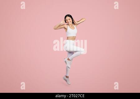 Donna eccitata con abbigliamento sportivo e cuffie wireless che saltano su sfondo rosa, foto a lunghezza intera, spazio libero Foto Stock