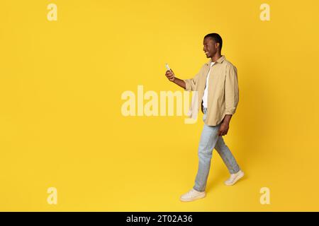 Uomo afro-americano millenario positivo in uso casuale telefono per chiacchierare, camminare Foto Stock