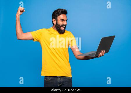 Gioioso uomo indiano di mezza età che tiene un portatile e celebra il successo, alzando il pugno stringato ed esclamando con gioia Foto Stock