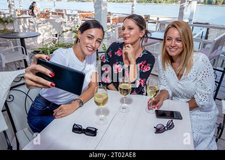Tre amiche allegri siedono a un tavolo con un bicchiere di vino bevendo, parlando e scattando selfie sulla terrazza di un ristorante sul fiume. Foto Stock