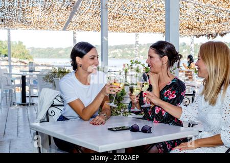 Tre amiche allegri siedono a un tavolo con un bicchiere di vino brindando, chiacchierando e ridendo sulla terrazza di un ristorante sul fiume. Foto Stock