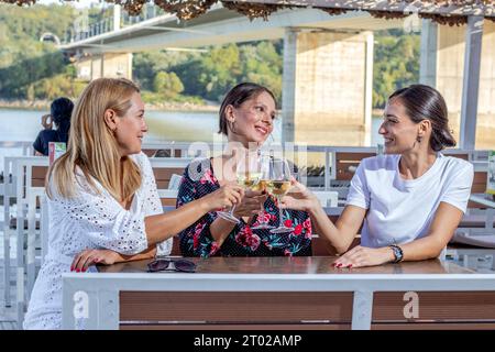 Tre amiche allegri siedono a un tavolo con un bicchiere di vino brindando, chiacchierando e ridendo sulla terrazza di un ristorante sul fiume. Foto Stock