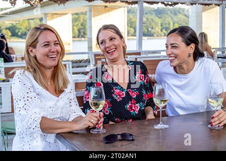 Tre amiche allegri siedono a un tavolo con un bicchiere di vino bevendo, chiacchierando e ridendo sulla terrazza di un ristorante sul fiume. Foto Stock