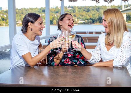 Tre amiche allegri siedono a un tavolo con un bicchiere di vino brindando, chiacchierando e ridendo sulla terrazza di un ristorante sul fiume. Foto Stock