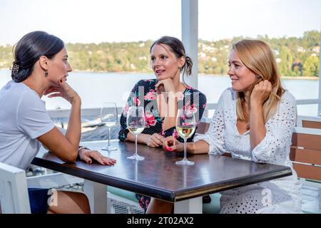 Tre amiche allegri siedono a un tavolo con un bicchiere di vino bevendo, chiacchierando e ridendo sulla terrazza di un ristorante sul fiume. Foto Stock