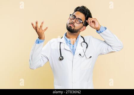 Non so cosa dire. Confuso giovane medico indiano cardiologo si sente imbarazzato per una domanda ambigua, avendo dubbi, nessuna risposta, incomprensibile. Un farmacista arabo su sfondo beige Foto Stock