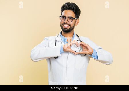 Medico indiano cardiologo uomo innamorato. Sorridente attraente farmacia araba fa gesto cardiaco dimostra segno d'amore esprime buoni sentimenti positivi isolato su sfondo beige Foto Stock