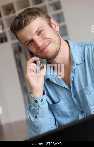 Bell'uomo parla al telefono e sorride. L'operatore risponde alla chiamata telefonica. Il ragazzo caucasico in camicia di denim è seduto in ufficio e parla al telefono cellulare. Emozioni positive. Servizio di supporto cordiale. Foto Stock