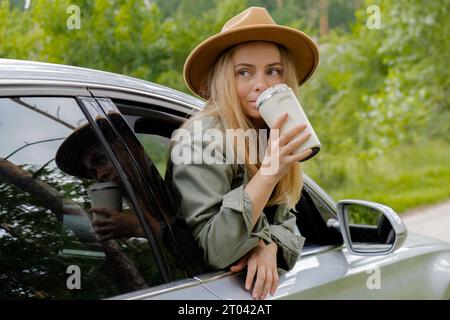 Donna bionda che esce dalla macchina del parabrezza e beve caffè o tè dalla tazza riutilizzabile. I giovani turisti esplorano i viaggi locali, trasformandoli in momenti autentici. Rifiuti riutilizzo riciclaggio zero rifiuti. Viaggiare in modo responsabile ridurre le emissioni di anidride carbonica stile di vita sostenibile Foto Stock