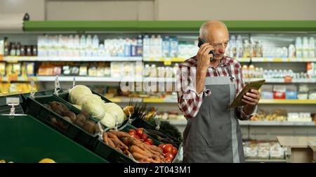 Anziano assistente di vendita maschile in una drogheria che utilizza un tablet digitale e parla al cellulare mentre fa un inventario Foto Stock