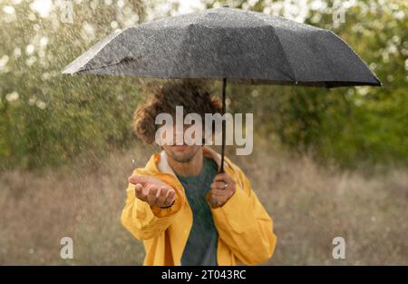 Giovane affascinante uomo arabo in impermeabile giallo con copertura per ombrello nera dalla pioggia nel parco Foto Stock