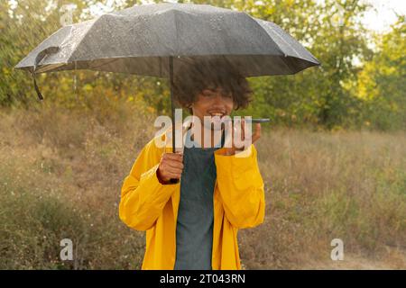 Giovane arabo che invia un messaggio vocale con lo smartphone che tiene un ombrello al parco Foto Stock