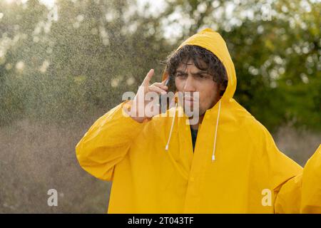 Uomo stanco con un impermeabile giallo e un ombrello di cattivo umore che parla con lo smartphone mentre cammina nel parco in una giornata di pioggia Foto Stock