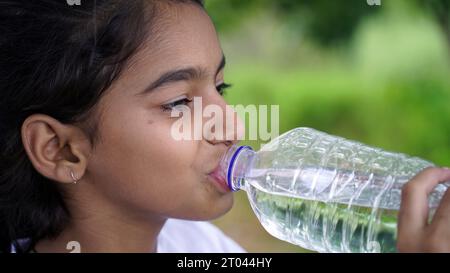 Simpatica e carina ragazza indiana con i capelli neri che beve acqua nel parco, tenendo una bottiglia di plastica e mostrando il pollice in alto. foto all'aperto Foto Stock