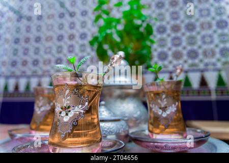 Tè moresco con menta, bevanda tradizionale marocchina in bicchieri di cristallo decorati, teiera argentata e ciotola da zucchero sullo sfondo Foto Stock