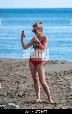 una giovane donna attraente che beve una lattina di birra con un bikini rosso che scatta una foto selfie su una spiaggia in vacanza a zante, grecia. Foto Stock