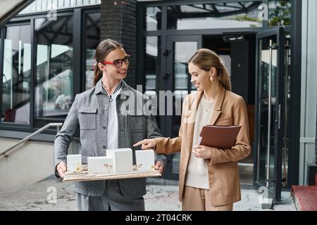 due colleghi sorridenti che discutono di un modello in scala che tiene documenti e sorride sinceramente, ufficio di design Foto Stock