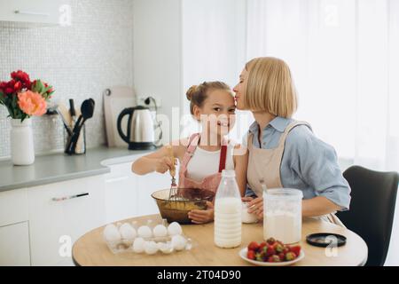 Madre e figlia stanno preparando impasto per pancake a tavola in una cucina moderna. La ragazza tiene una frusta, mescola le uova in una ciotola, prepara l'impasto con la h. Foto Stock