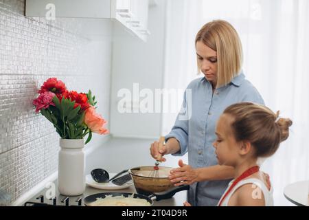 Una giovane madre e sua figlia cucinano i pancake insieme per colazione. La madre insegna alla figlia a cucinare. Foto Stock