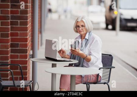 Anziana donna d'affari dai capelli grigi seduta in una caffetteria in strada con telefono cellulare e computer portatile, donna anziana che lavora utilizzando tecnologie e gadget. Foto Stock