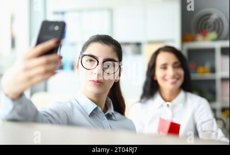 La giovane donna in occhiali tiene il telefono davanti al viso e sorride ai colleghi in ufficio Foto Stock